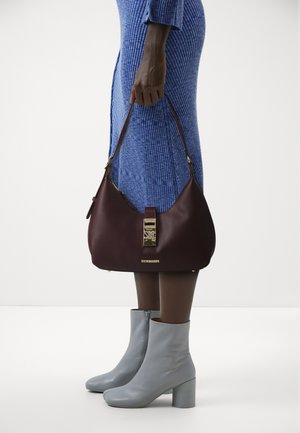 Borsa a tracolla in pelle marrone con dettagli dorati, dotata di chiusura e logo. Abbinata a stivali alla caviglia grigio chiaro e a un vestito blu strutturato.