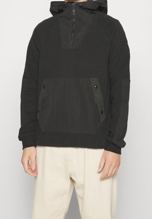 Sweat à capuche noir à enfiler avec fermeture éclair devant, poche kangourou à boutons et capuche avec cordon, porté avec un pantalon beige clair.