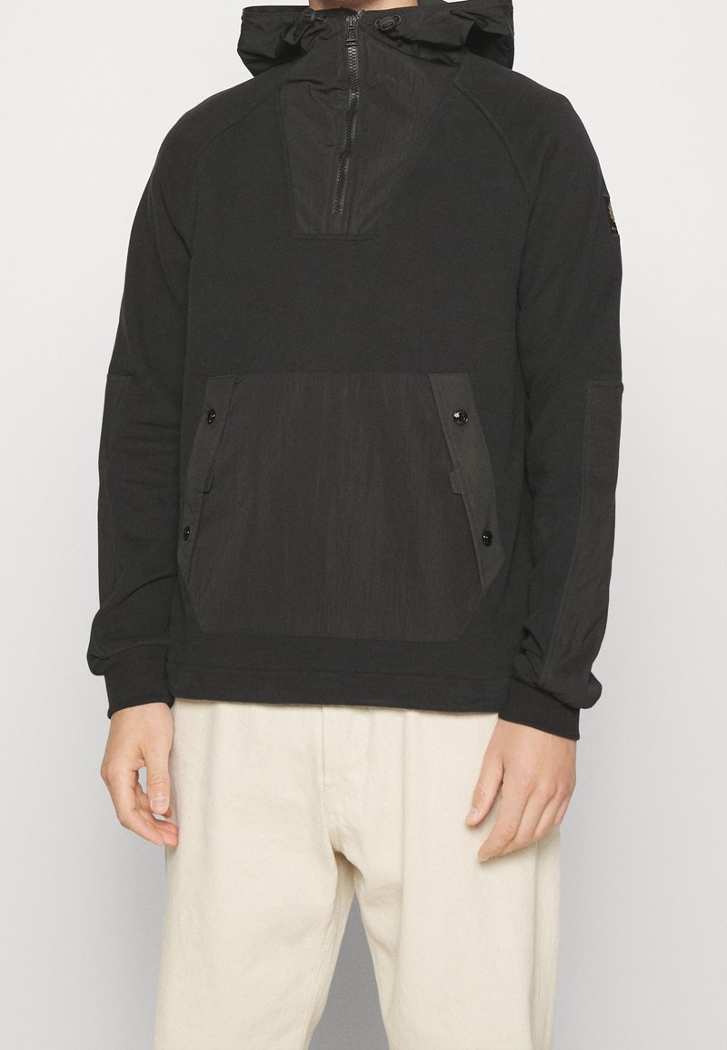 Sweat à capuche noir à enfiler avec fermeture éclair devant, poche kangourou à boutons et capuche avec cordon, porté avec un pantalon beige clair.