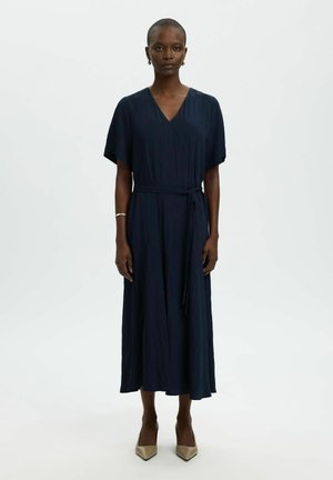 Mujer de pie vestida con un vestido midi azul marino con escote en V, mangas cortas y un cinturón anudado en la cintura, combinado con tacones beige puntiagudos.