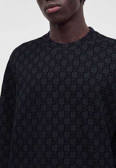 Gros plan d'une personne portant une chemise noire à motifs avec un col rond haut et des formes abstraites répétitives.