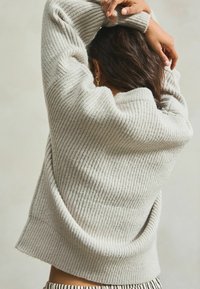 Lichtgrijze geribde trui met een losse pasvorm, brede ronde hals en textuurstof. De achterkant is iets langer en is een gestreepte onderkant zichtbaar.