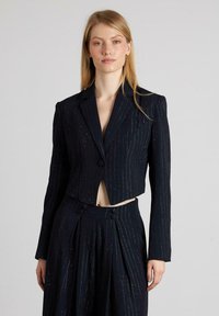 Giacca blu navy tagliata con righe sottili e leggera lucentezza. Presenta un colletto a scollatura e chiusura con un solo bottone; maniche lunghe.