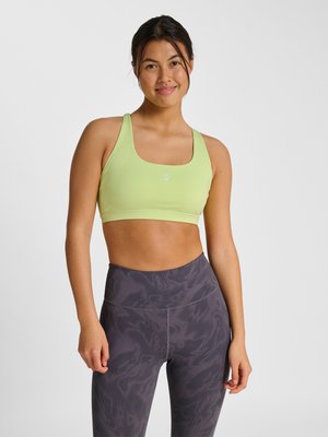 Kvinde iført en lysegrøn sports-bh og grå leggings med højtalje og mønster, stående afslappet mod en neutral baggrund.