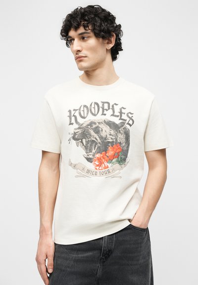 Νέος άνδρας με σκούρα σγουρά μαλλιά που φοράει ένα εκρού μπλουζάκι με γραφικό σχέδιο ενός αγριεμένου πάνθηρα και κόκκινα λουλούδια, καθώς και μαύρο τζιν παντελόνι.