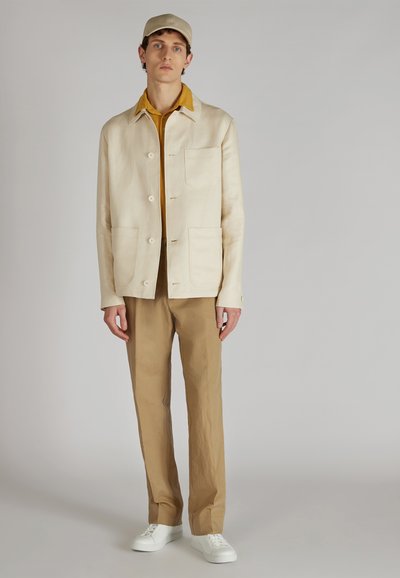 Veste en lin beige avec de grandes poches, associée à un polo moutarde et un pantalon marron clair. Des baskets blanches complètent l'ensemble.