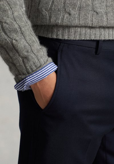 Person wearing a gray cable-knit sweater with blue and white striped shirt cuffs showing, hand partially in the front pocket of dark pants.