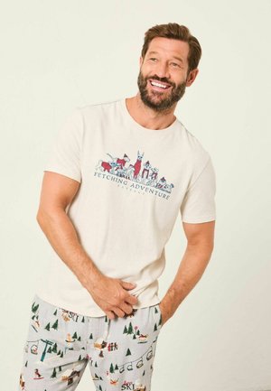 Helles beige T-Shirt mit einem Grafikmotiv "Fesselndes Abenteuer" von Tieren. Kombiniert mit gemusterten Schlafanzughosen, die Skimotive und Kiefern zeigen.