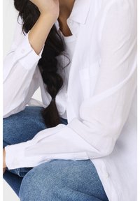 Valkoinen, pitkähihaista nappipaitaa, jossa on teksturoitu pellavamateriaali. Malli istuu sinisissä farkuissa, mikä näkyy napin yksityiskohtana hihoissa.