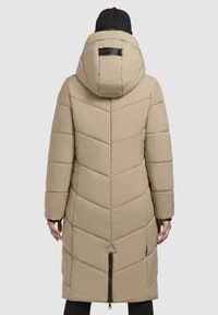 Lang beige pufferjakke med et chevron-quiltet mønster, stor hætte og sort lynlås lukning i bunden. Branding på ryggen.