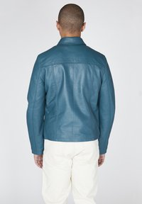 Giacca di pelle verde acqua con colletto classico e design aderente, caratterizzata da dettagli di cucitura sulla schiena e maniche lunghe. Pantaloni bianchi semplici.
