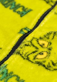 Tessuto in pile giallo brillante con una cerniera nera, raffigurante una grafica di una testa di serpente a sonagli e testo parzialmente visibile.