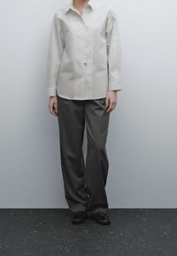 Persona che indossa una camicia a righe bianche a maniche lunghe, pantaloni grigi larghi e mocassini neri, in piedi contro un muro grigio chiaro semplice.