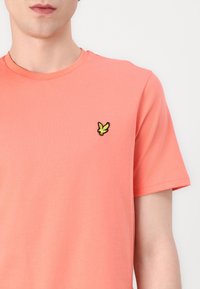 Korallfärgad T-shirt i mjuk bomull, med rund halsringning och korta ärmar. En liten, svart och gul broderad fågel-logotyp på bröstet.
