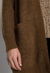 Brązowy sweter kardigan z dzianiny o teksturowanym wykończeniu i ściągaczami na rękawach, z naszytą kieszenią z przodu i luźnym krojem.