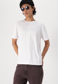 Bijela pamučna majica s logotipom "Hollister" u svijetlom printu, u kombinaciji s tamno smešnim trapericama i okruglim naočalama za sunce. Jednostavan, ležeran dizajn.