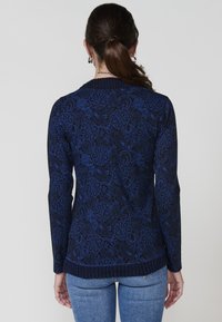 Dunkelblauer Strickpullover mit floralem Paisleymuster, langen Ärmeln, geripptem Saum und glatter Textur. Trägt dazu eine hellblaue Jeans.