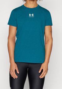 Person, die ein türkisfarbenes Under Armour T-Shirt und schwarze Leggings trägt, sichtbar von den Schultern bis zur Mitte der Oberschenkel, mit sichtbaren Tätowierungen auf dem Arm.