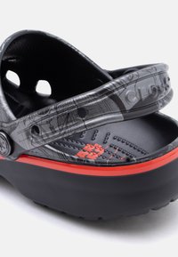 Μαύρο και γκρι σανδάλι Crocs με κόκκινο λογότυπο και ιμάντα με λεπτομερή σχέδιο, τοποθετημένο σε γωνία για να δείχνει τη φτέρνα και το πλάι.