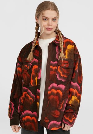 Giacca camicia in fleece marrone con ampi motivi floreali multicolori in arancione, rosa e giallo. Dotata di chiusure a bottone e due tasche sul petto.