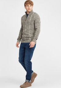 Sivý textúrovaný zipsový sveter s vysokým golierom, skombinovaný s modrými džínsami a svetlohnedými semišovými topánkami. Drevený detail na ťaháku zipsu.