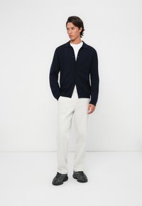Cardigan bleumarin cu guler și închidere prin nasturi, purtat peste o cămașă albă, asortat cu pantaloni de culoare deschisă și adidași negri.