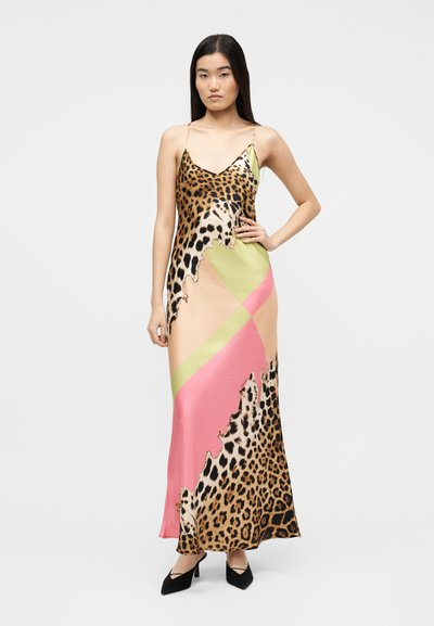 Vrouw staat met hand op heup en draagt een lange slipjurk met luipaardprint en roze-groene kleurvlakken, gecombineerd met zwarte hakken.