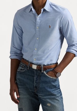 Homme en chemise bleu clair à boutons avec manches retroussées, ceinture en cuir marron, jean bleu foncé et montre-bracelet marron, main dans la poche.