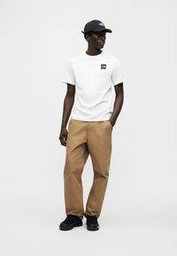 Λ ευκό κοντομάνικο T-shirt με μαύρο λογότυπο, συνδυασμένο με καφέ cargo παντελόνι και μαύρα αθλητικά παπούτσια, ολοκληρωμένο με μαύρο καπέλο μπέιζμπολ.