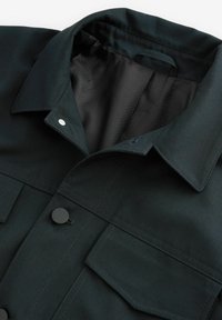 Mørkegrøn jakke med struktureret krave, to frontlommer og trykknapper. Stofoverfladen er glat med et subtilt ribbet mønster.