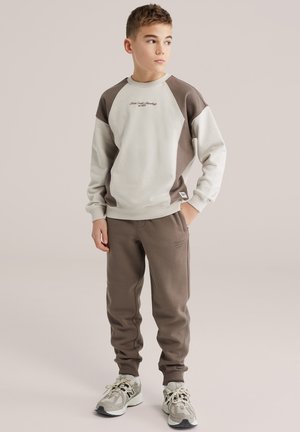 Fant z rokami v žepih, oblečen v bež in rjavo jopico, rjave športne hlače ter sive superge, gleda vstran.