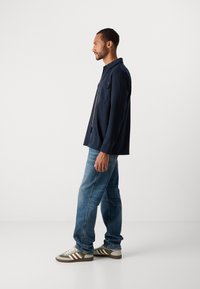 Man standing sideways wearing a navy button-up shirt, blue jeans, and beige and olive Adidas sneakers against a plain white background.