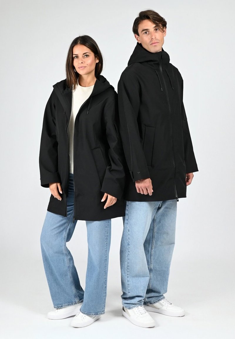 Giubbotti impermeabili neri con cappuccio, con chiusura frontale con cerniera e tasche laterali, indossati sopra jeans azzurri casual e scarpe bianche.