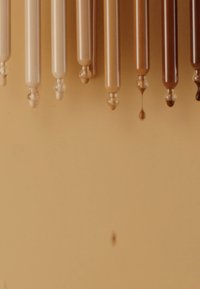 Sett med åtte lange, sylindriske rør i varierende nyanser av beige til brun, med glansfulle overflater og runde hetter, som drypper farge mot en beige bakgrunn.