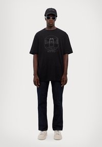 Črna prevelika majica s grafičnim dizajnom v sivi barvi, v kombinaciji s temnimi kavbojkami in belimi supergami. Dodatki vključujejo čepico in sončna očala v črni barvi.