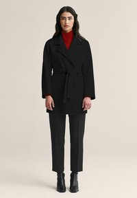 Musta vyöllä varustettu takki, jossa on lovikaulus, puettu punaisen poolopaidan päälle, yhdistetty mustiin housuihin ja nilkkureihin, luoden yksivärisen ilmeen.