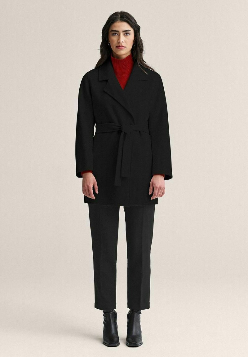 Musta vyöllä varustettu takki, jossa on lovikaulus, puettu punaisen poolopaidan päälle, yhdistetty mustiin housuihin ja nilkkureihin, luoden yksivärisen ilmeen.