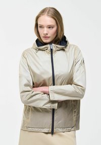 Mlada ženska z blond lasmi v bež jakni s kapuco, prekrižanimi rokami, rahlo obrnjena v stran z nevtralnim izrazom na obrazu.