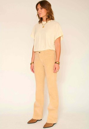 Pantalons en velours côtelé beige clair évasés avec une finition texturée, associés à un haut ample crème, portés avec des ballerines marron à bout pointu.