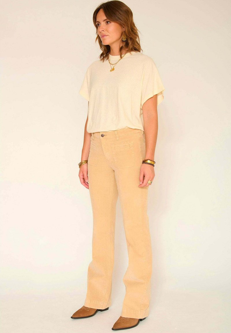 Pantalons en velours côtelé beige clair évasés avec une finition texturée, associés à un haut ample crème, portés avec des ballerines marron à bout pointu.