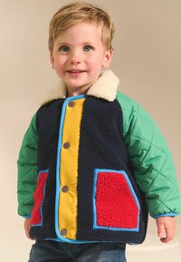 Veste colorée avec un corps en polaire bleu marine, des manches matelassées vertes, une patte de boutonnage jaune et des poches rouges, dotée d'un col en fourrure blanche.