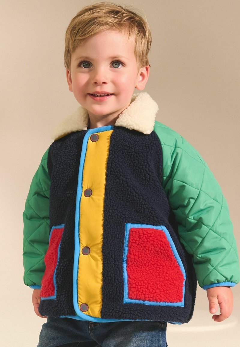 Veste d'hiver multicolore avec un design bleu marine, vert et jaune ; présente une texture pelucheuse, un col en faux fourrure et des poches contrastantes.