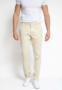 Hombre con pantalones beis ajustados, camiseta blanca y zapatillas blancas, de pie contra un fondo blanco liso.