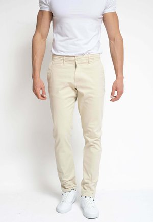 Hombre con pantalones beis ajustados, camiseta blanca y zapatillas blancas, de pie contra un fondo blanco liso.