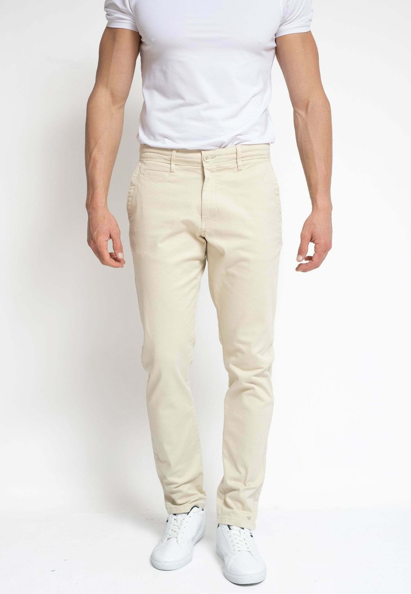 Hombre con pantalones beis ajustados, camiseta blanca y zapatillas blancas, de pie contra un fondo blanco liso.