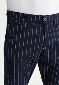 Marinblå randiga byxor i bomull, med vita vertikala ränder, knäppning med knapp och sidofickor för funktionalitet.