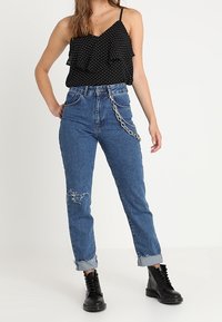 Černý top na ramínkách s puntíky a volánkovým detailem. Modré džíny z denimu s obnošenými akcenty a ohrnutými lemy. Černé boty s tkaničkami až ke kotníkům.