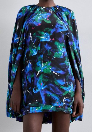 Model draagt een losse, knielange jurk met een abstract bloemenpatroon in blauw, groen en zwart en wijd uitlopende mouwen tegen een effen achtergrond.