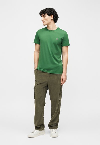 Νέος άνδρας με σγουρά μαλλιά στέκεται με τα χέρια στις τσέπες, φορώντας πράσινο κοντομάνικο πουκάμισο, χακί παντελόνια cargo και λευκά αθλητικά παπούτσια, κοιτάζοντας αριστερά.