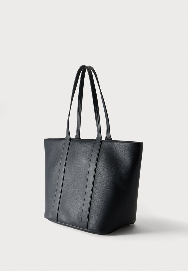 Borsa tote in pelle nera con due manici lunghi e una texture liscia, posizionata in piedi su uno sfondo bianco uniforme.